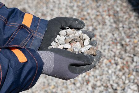 Nahaufnahme von behandschuhten Händen in Arbeitskleidung, die eine Menge rezyklierte Gesteinskörnung halten. Im Hintergrund ist eine unscharfe Fläche aus ähnlichem Kies zu sehen.