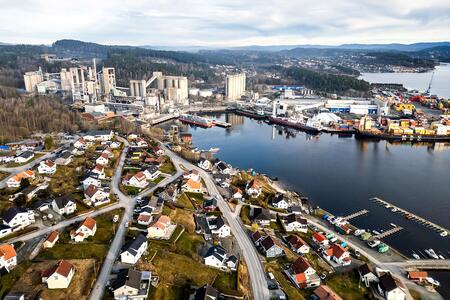 Luftaufnahme von Brevik in Norwegen mit dicht bebauten Wohnhäusern im Vordergrund, großem Zementwerk auf der linken Seite sowie einem Containerhafen rechts am Wasser. Im Hintergrund bewaldete Hügel und ein See.