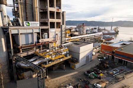 Zementwerk Brevik im Vordergrund mit Heidelberg Materials-Logo und technischer Ausrüstung. Im Hintergrund sind ein Hafen, Kräne und ein Fluss zu sehen. Zahlreiche Fahrzeuge und Container befinden sich im unteren rechten Bereich.