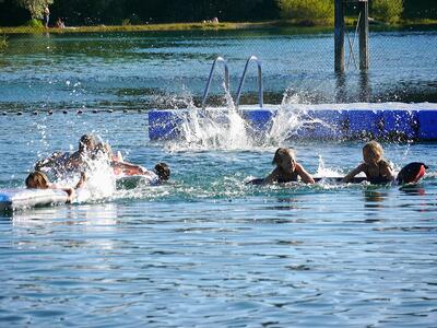 Kinder schwimmen in einem See. Im Hintergrund befinden sich ein Steg, eine Leiter und Bojen, die den Schwimmbereich abgrenzen. Spritzendes Wasser zeigt aktive Bewegung.