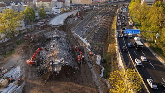 Luftaufnahme einer Baustelle neben einer Autobahn über Eisenbahnschienen in einer Stadt. Im linken Bereich ist ein großer Erdhügel mit Baggern und Bauarbeitern zu sehen. Rechts verlaufen dicht befahrene Fahrspuren der Autobahn parallel zu Gleisen. Im Hintergrund sind Stadtgebäude und Bäume im Abendlicht erkennbar.