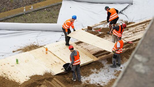 Fünf Bauarbeiter in orangefarbenen Sicherheitswesten verlegen Holzplatten über die mit weißer Plane abgedeckten Gleise; zwei Arbeiter halten eine Platte, die sie positionieren.