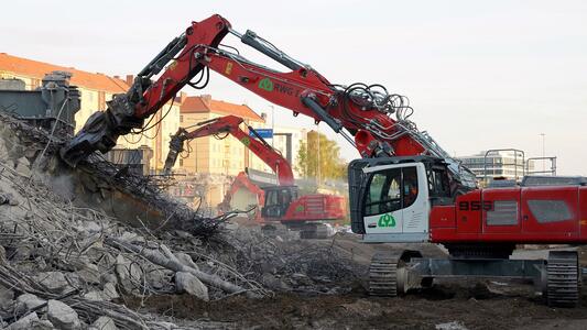 Zwei rot-weiße Abrissbagger entfernen Trümmer einer abgerissenen Brücke auf einer Baustelle. Im Hintergrund sind Gebäude sichtbar.