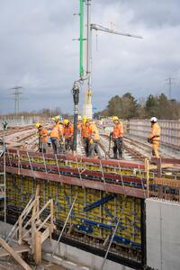 Bauarbeiter in orangefarbener Sicherheitskleidung arbeiten auf einer Brückenbaustelle. Sichtbar sind eine Betonpumpe, die Beton auf die Brücke gießt, und ein Kran im Hintergrund. Umgeben von freiem Himmel und Strommasten in der Ferne.