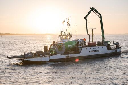 Fähre mit Betonmischer auf Deck fährt über das Wasser bei Sonnenuntergang, das Wasser reflektiert das Licht; im Hintergrund schwache Konturen einer Uferlinie sichtbar.