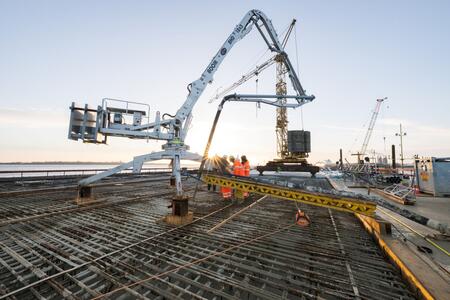 Baustelle mit Betonpumpe und Bauarbeitern, die Beton auf eine Stahlbewehrung gießen; im Hintergrund Kräne und Sonnenaufgang, zentrale Betonierarbeiten auf Brückenfundament.