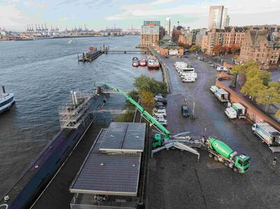 Eine Betonpumpe pumpt Beton in ein U-Boot am Ufer eines Flusses. Links der Hafen mit Booten, rechts eine Straße mit parkenden Fahrzeugen und Gebäuden im Hintergrund. Im Vordergrund eine grüne Betonpumpe, die in das Schiff ragt.