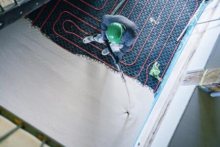 Arbeiter mit grünem Helm trägt Estrich auf Fußbodenheizung auf; rote Heizschlangen sind teils sichtbar, Estrich wird gleichmäßig verteilt, Aufnahme aus Vogelperspektive.