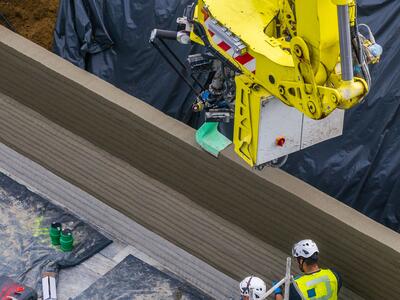 Gelber 3D-Drucker beim Schichten von Druckbeton für den Bau einer Wand; zwei Bauarbeiter mit Schutzhelmen beobachten und sichern die Arbeiten unten rechts. Im Hintergrund schwarze Abdeckfolie und Baustellenmaterialien.