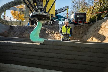 Ein Bauarbeiter überwacht einen 3D-Drucker, der Schichten aus Beton aufträgt, um einen Keller zu bauen. Im Hintergrund sind Baustellenfahrzeuge und herbstlich gefärbte Bäume sichtbar.