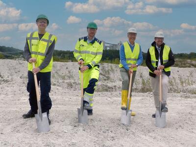 Vier Personen mit Schutzkleidung halten Spaten auf einer Baustelle.