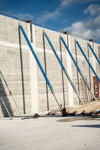 Große 3D-gedruckte Betonwand mit blauen Stützen auf einer Baustelle bei sonnigem Wetter.
