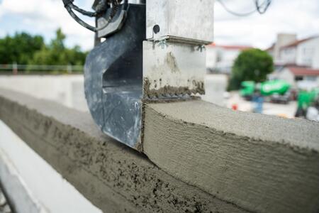 Druckbeton wird mithilfe eines Druckers präzise aufgetragen, im Hintergrund ist eine Baustelle sichtbar.