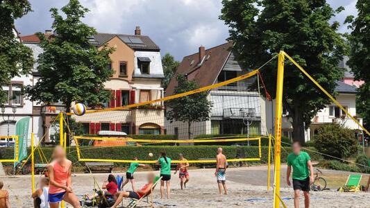Menschen spielen Beachvolleyball auf einem Sandplatz, umgeben von Bäumen und Häusern.