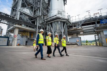 Personengruppe mit Schutzhelmen und Warnwesten geht vor einer industriellen Anlage.