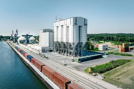 Industrieanlage mit mehreren Silos und Schienenverkehrsanbindung an einem Flussufer.