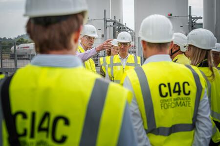 Personengruppe in Sicherheitswesten und Helmen auf einer Industrieanlage, ein Mann zeigt gestikulierend auf etwas.