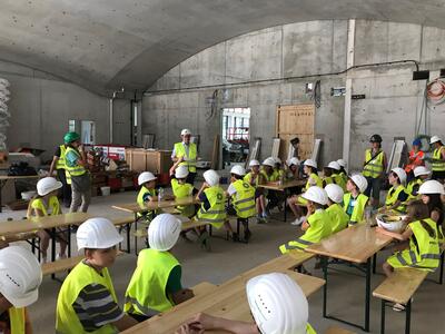 Kinder mit Schutzhelmen und Warnwesten hören in einer Baustellenumgebung einer Präsentation zu.