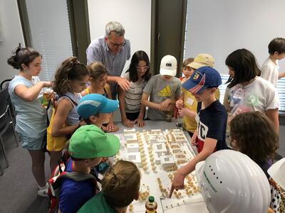 Eine Gruppe von Kindern und ein erwachsener Mann stehen um ein Architekturmodell auf einem Tisch. Die Kinder tragen Freizeitkleidung und Mützen, einige berühren oder betrachten das Modell, während der Mann etwas erklärt. Im Hintergrund sind große, lichtdurchflutete Fenster zu sehen.