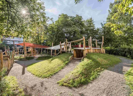 Ein Spielplatz im Freien in einer üppig grünen Umgebung mit Bäumen und Sonnenlicht, das durch das Laub filtert. Der Spielplatz umfasst Holzstrukturen wie ein Spielhaus, Brücken und Plattformen sowie grasbewachsene Hügel.