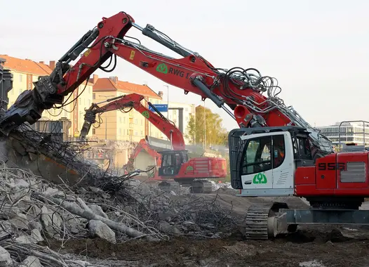 Zwei rot-weiße Abrissbagger entfernen Trümmer einer abgerissenen Brücke auf einer Baustelle. Im Hintergrund sind Gebäude sichtbar.