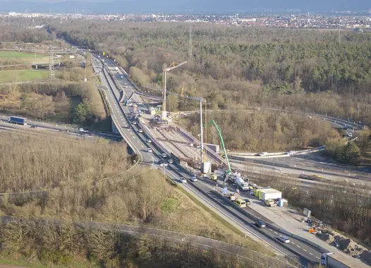 Luftaufnahme einer Autobahnbaustelle: Ein mehrspuriger Autobahnabschnitt mit laufenden Brückenarbeiten in der Bildmitte. Zwei große Kräne stehen auf der Baustelle, umgeben von Fahrzeugen und Baumaschinen. Verkehr fließt rechts und links um die Baustelle herum. Im Hintergrund sind Wälder, landwirtschaftliche Flächen und eine Stadt zu sehen.