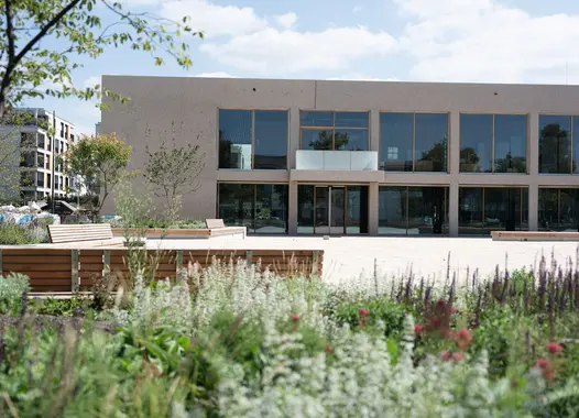 Frontansicht des zweigeschossigen Bürgerhauses mit großen Fensterflächen, vorgelagerter Platzgestaltung und bepflanzten Aufenthaltsbereichen.