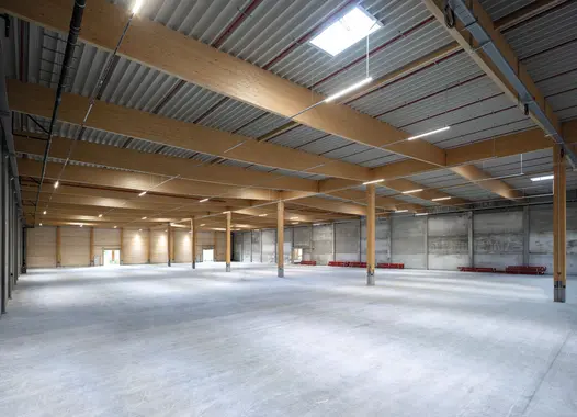 Große, leere Lagerhalle mit Holzstützen, Sichtbetonwänden und einer Stahlträgern und Lichtöffnungen. Der Boden ist betoniert und unbelegt; auf der rechten Seite liegen rote Bauelemente. Im Hintergrund befinden sich mehrere Tore. Tageslicht fällt durch Öffnungen im Dach ein.