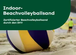 Nahaufnahme eines Volleyballs im Sand in einer Indoor-Beachvolleyballhalle. Im Hintergrund sind unscharf zwei Personen beim Spielen vor einem gelben Netz zu sehen. Links oben befindet sich ein dunkelgrünes Banner mit der Aufschrift „Indoor-Beachvolleyballsand – Zertifizierter Beachvolleyballsand durch den DVV“.